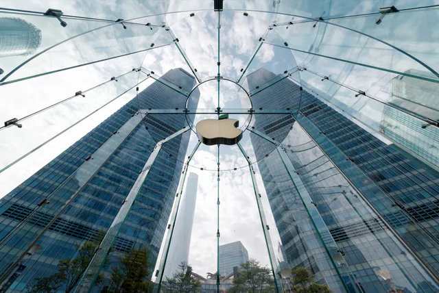 Looking up through the glass cylinder entrance of the Apple Store in Pudong, Shanghai, with the Apple logo suspended at the center and skyscrapers rising behind it