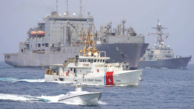 A cargo ship and U.S. naval vessels transit the Strait of Hormuz behind an unmanned surface vessel.