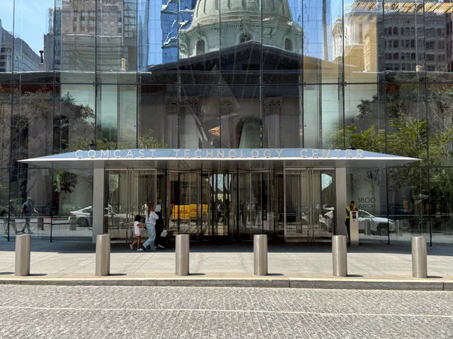 Glass-walled street-level entrance with a metal canopy reading 'COMCAST TECHNOLOGY CENTER,' the dome of Philadelphia City Hall reflected in the facade, pedestrians walking past.