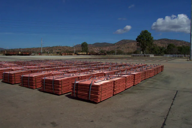 Rows of strapped bundles of reddish copper cathode sheets stacked in an outdoor shipping yard under a blue sky, with rail cars and hills in the background.