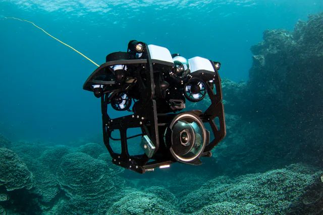 A black-framed remotely operated underwater vehicle with thrusters and camera domes hovers over a coral reef, a thin yellow control tether trailing off to the surface.