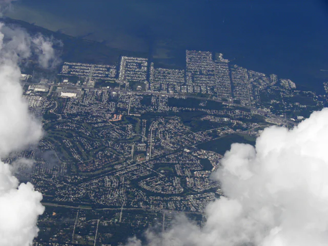 High-altitude aerial photograph of dense single-family-home subdivisions and canals along the Florida Gulf coast, partly framed by cumulus clouds.