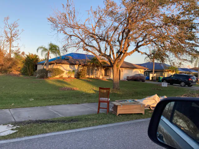 A single-story suburban Florida house with a blue tarp over its hurricane-damaged roof and damaged furniture piled at the curb, with neighboring homes visible down the street.
