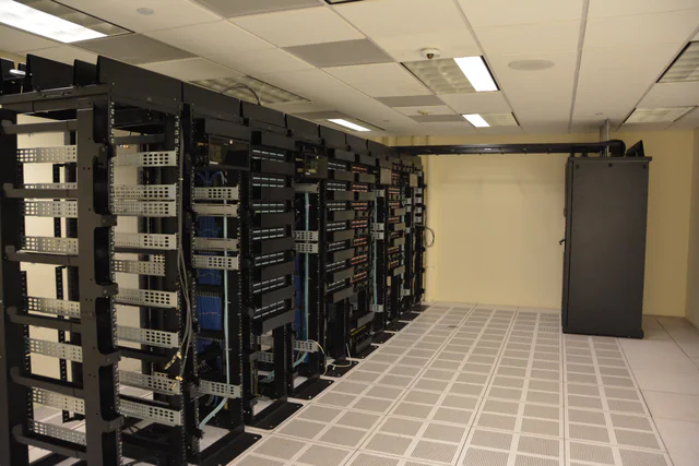 Long aisle between tall racks of networking and server equipment inside a data center, lit by blue and white indicator lights.