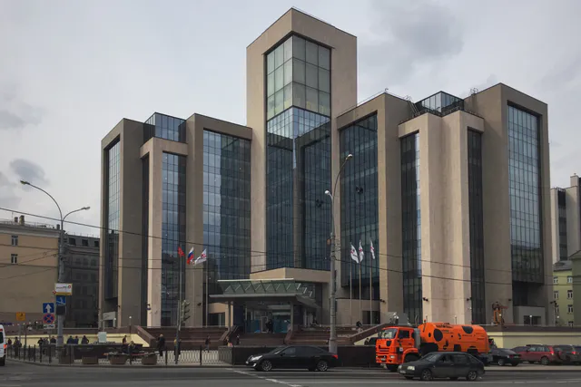 The Lukoil corporate headquarters building in Moscow, a large modern stone-and-glass office complex with Russian flags out front and cars passing on the street below.