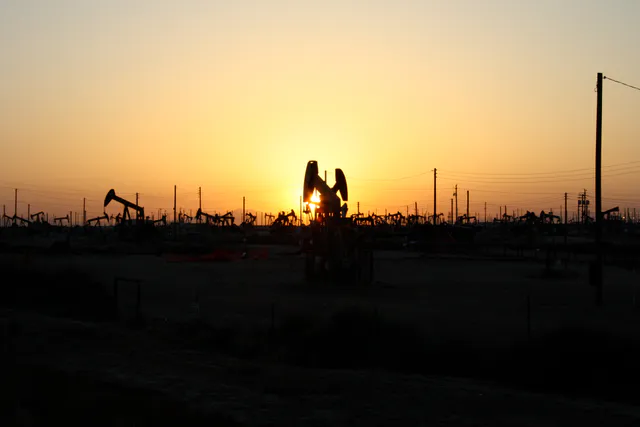 Rows of oil pumpjacks silhouetted against an orange sunset sky on the Lost Hills oil field in California.