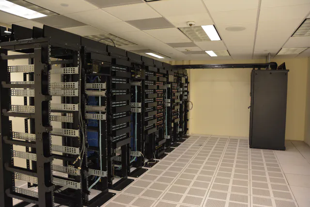 Long aisle of illuminated server racks lined with cabling inside a data center, viewed down the corridor between two rows of equipment.