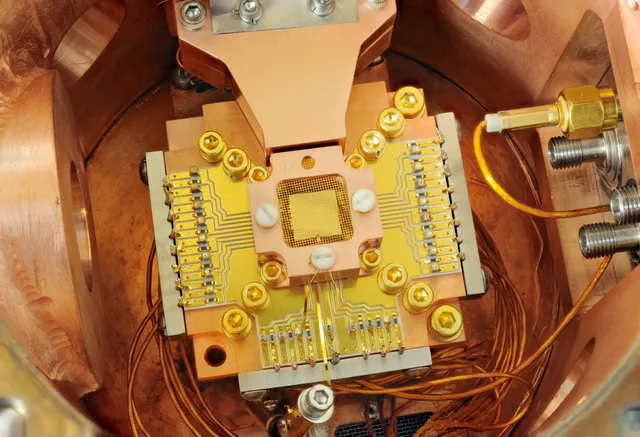 Close-up of a NIST trapped-ion quantum computing apparatus showing a gold ion-trap chip surrounded by copper enclosure and a gold wire mesh used to manipulate beryllium ions.