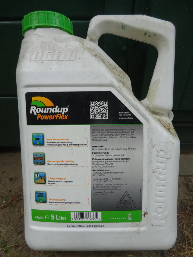 A white five-liter plastic canister of Roundup PowerFlex herbicide with green cap and Monsanto branding, photographed outdoors against a dark green door.