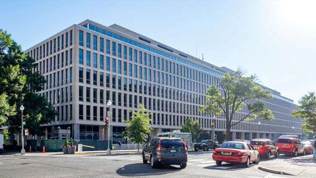 Exterior of the U.S. Department of Education headquarters, a long modernist office building in Washington, DC, on a sunny day with traffic passing in front.