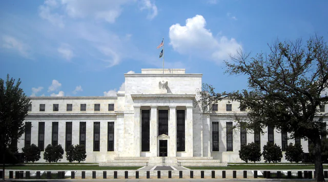 The neoclassical white marble facade of the Federal Reserve's Eccles Building in Washington, D.C., seen from the street under a clear sky.