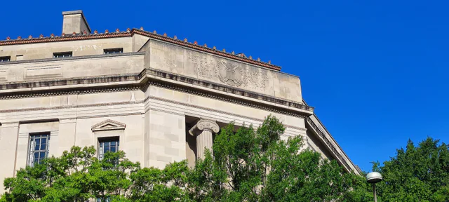 Upward view of the limestone Robert F. Kennedy Department of Justice Building under a clear blue sky, showing an Ionic column and a carved frieze reading JUSTICE against green treetops.