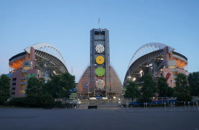 Lumen Field in Seattle's Pioneer Square, photographed from its north side at dusk with floodlights on and the downtown skyline behind it.