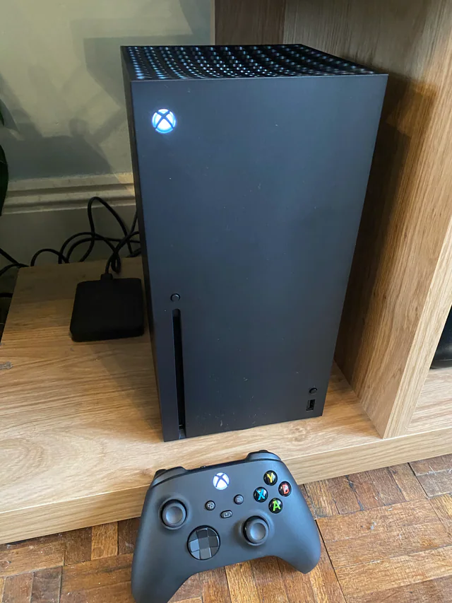 A black Xbox Series X console lying on a wooden surface next to a black Xbox wireless controller, both with illuminated power logos.